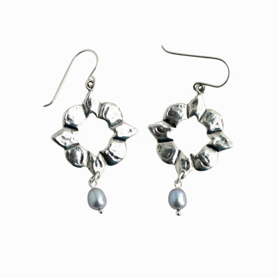 Long sterling silver earrings with bezel set pearls dangle from wood. 