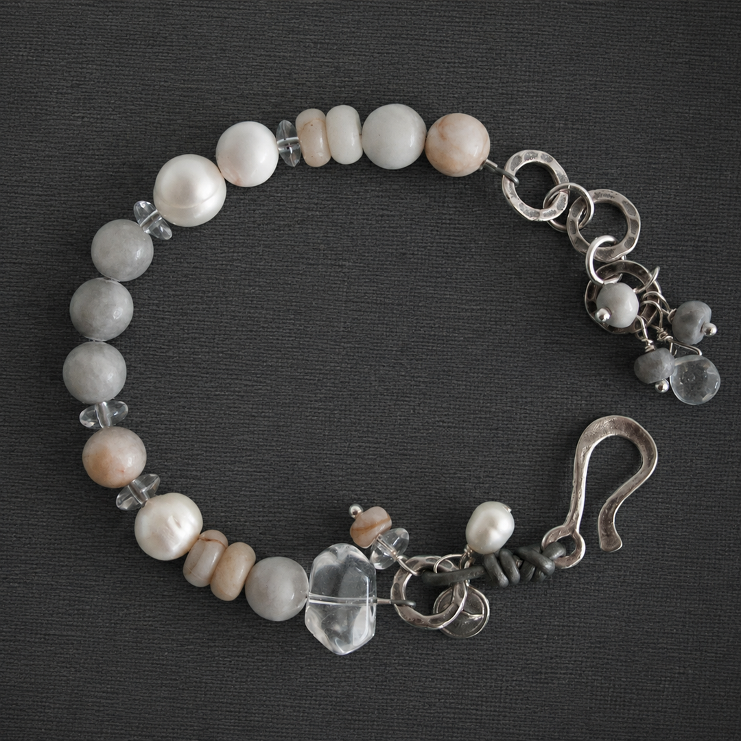 On the Rocks Bracelet with white pearls, gray and cream stones, clear quartz accents, silver links, charms, and hook clasp on a dark textured background.