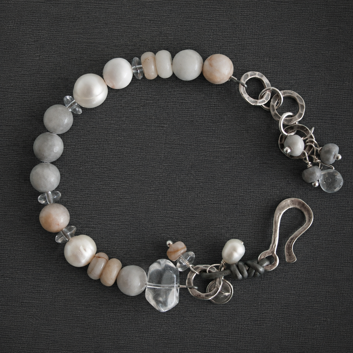On the Rocks Bracelet with white pearls, gray and cream stones, clear quartz accents, silver links, charms, and hook clasp on a dark textured background.