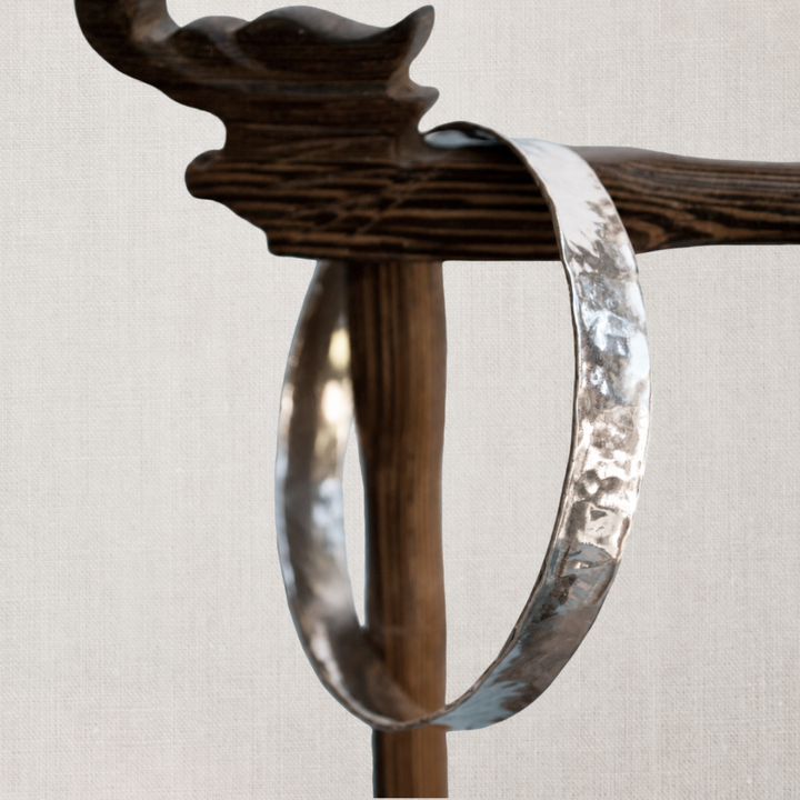 Modern Hammered sterling silver bangle bracelet displayed on a dark wood jewelry stand against a soft white linen background.