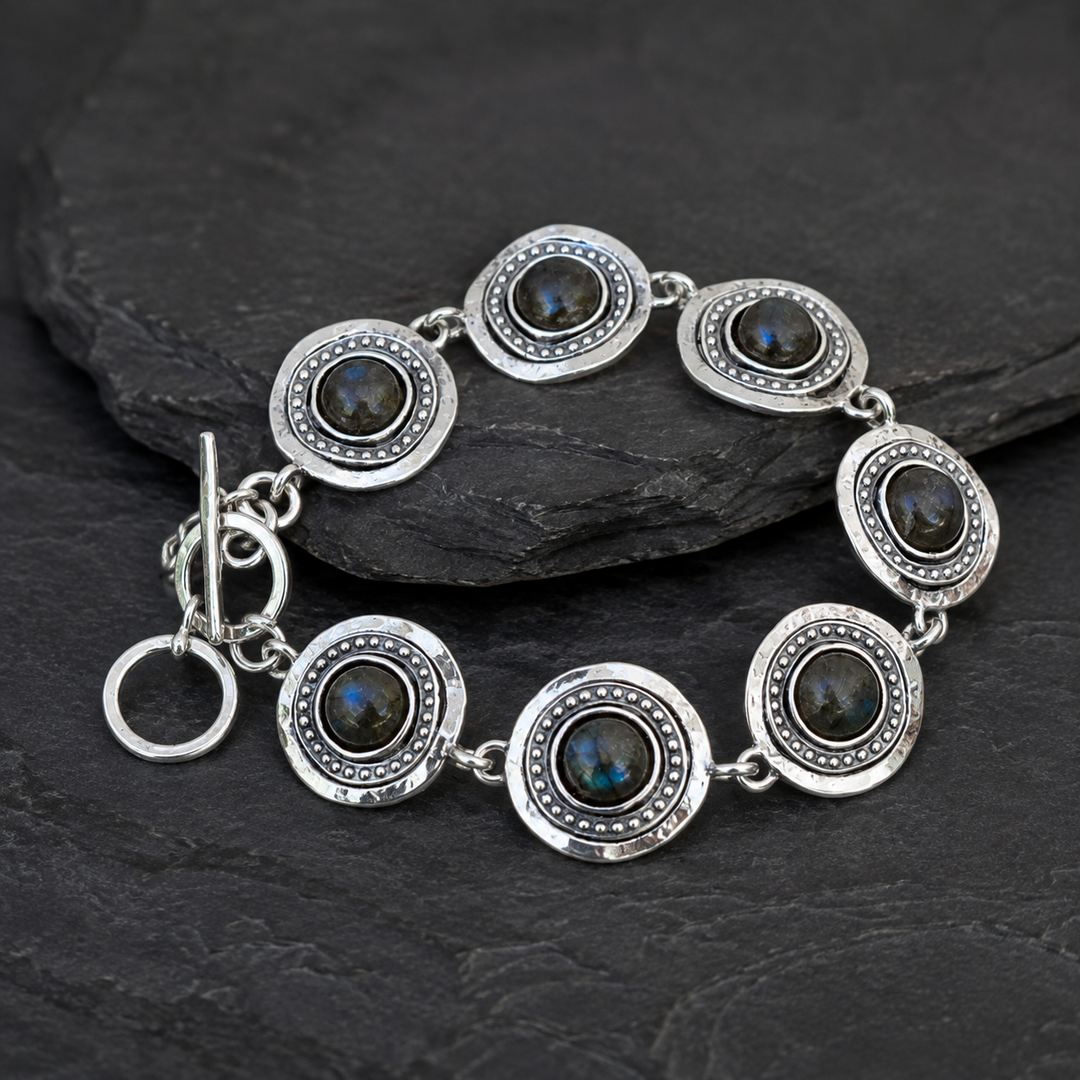 A sterling silver bracelet with seven oval links, each featuring a dark labradorite stone in the center and a beaded border, displayed on a dark slate surface.