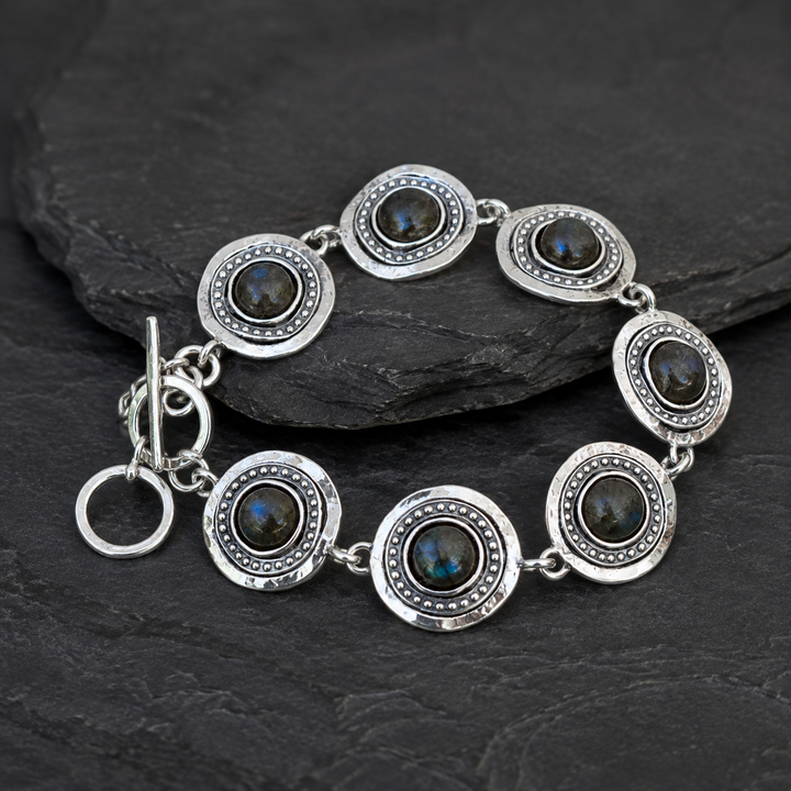 A sterling silver bracelet with seven oval links, each featuring a dark labradorite stone in the center and a beaded border, displayed on a dark slate surface.