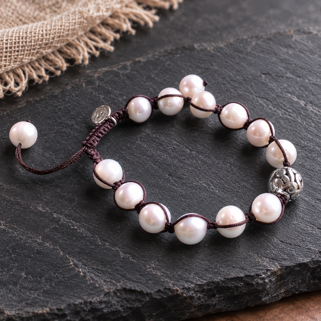 A beaded bracelet with white freshwater pearls threaded on a dark brown cord, featuring a silver-toned decorative accent bead and adjustable sliding knot closure, displayed on a dark slate surface.
