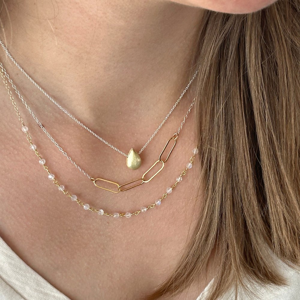 A close-up image of a person wearing a layered necklace with gold-colored links and a rainbow moonstone pendant.