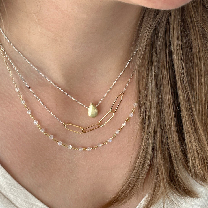 A close-up image of a person wearing a layered necklace with gold-colored links and a rainbow moonstone pendant.