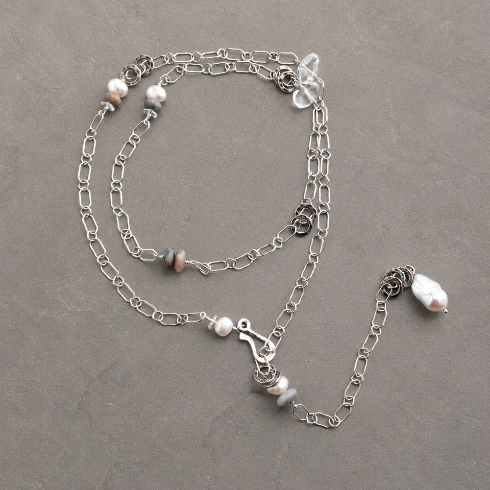 On the Rocks Necklace with sterling silver links, freshwater pearls, gray stones, and clear quartz accents arranged on a soft gray-brown slate background.