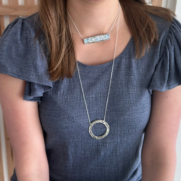 A person wearing a gray-blue shirt is showcasing a long silver necklace with a circular pendant bearing the words "love," "hope," "belief," and "dreams."