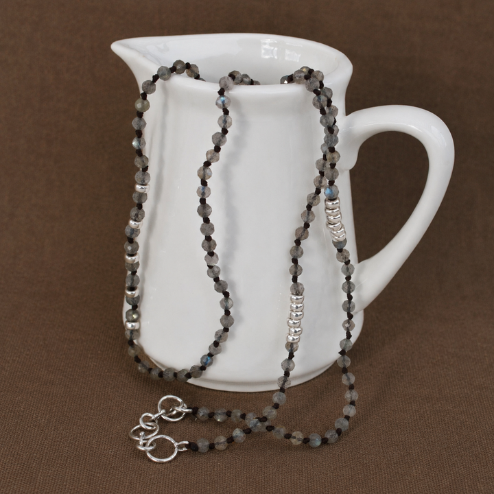 Streamside Labradorite Necklace with faceted gray labradorite beads and sterling silver accents draped over a white ceramic pitcher on a warm brown background.