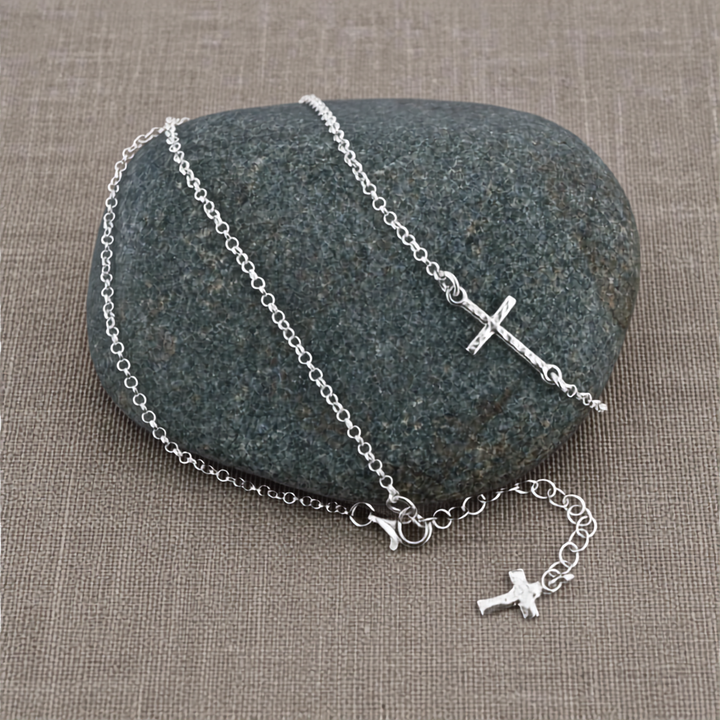 Sterling Silver Sideways Cross Necklace displayed on a dark stone with the extender chain and small cross charm visible on a neutral linen background.