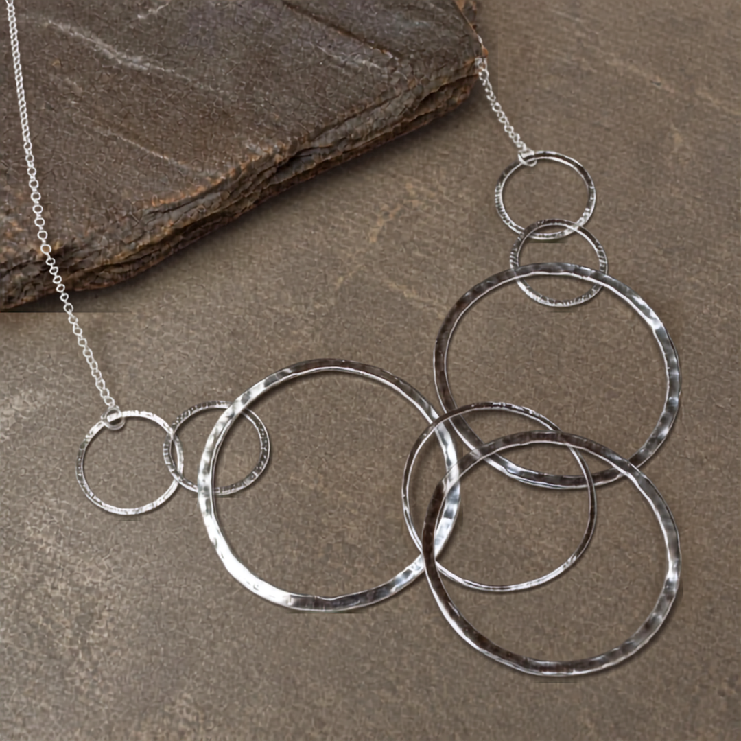 Our sterling silver Around We Go necklace, layered with hammered circular rings of varying sizes on a chain, is displayed on a textured brown stone surface.