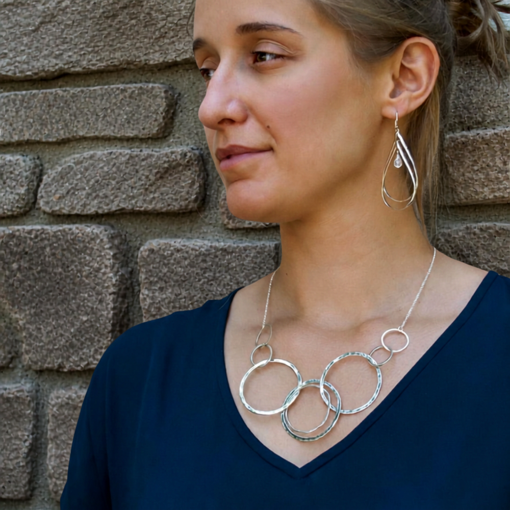 The Around We Go silver statement necklace with interlocking textured circles is worn by a model, who is photographed against a rustic stone wall.