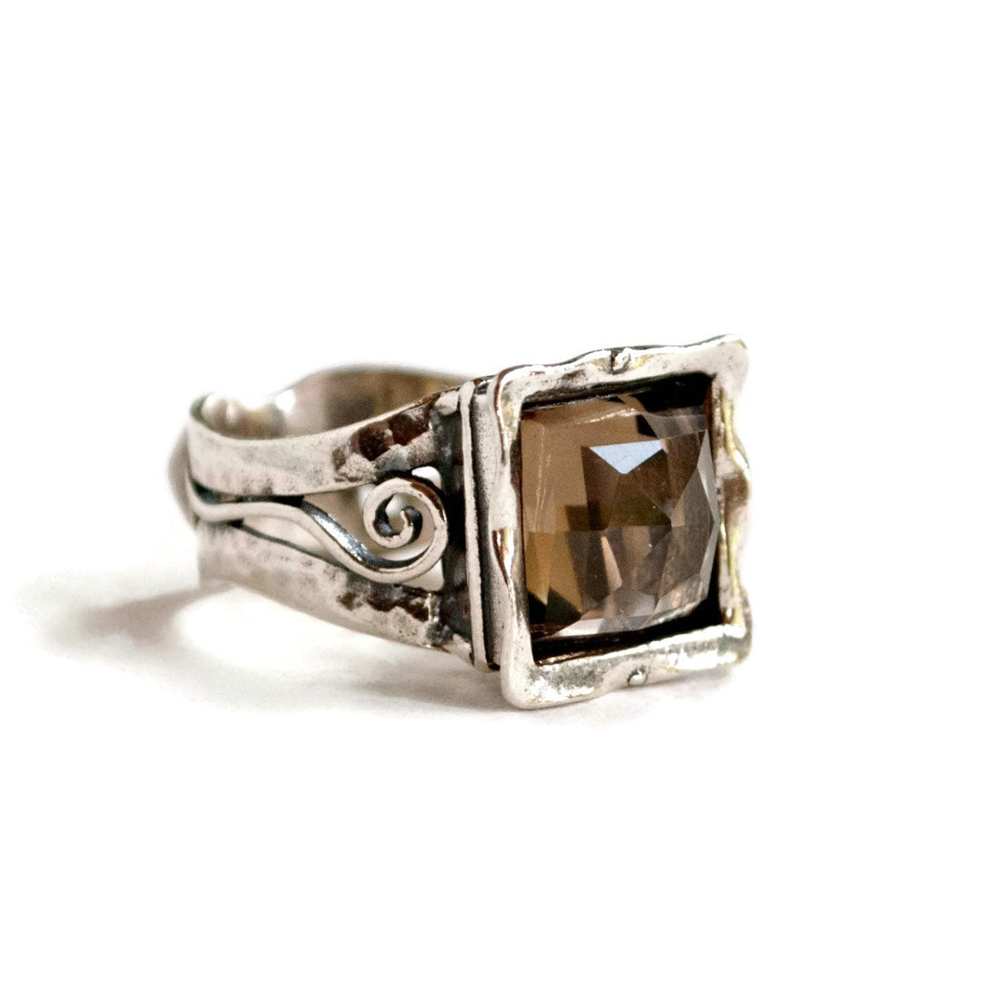A sterling silver ring with a faceted smoky quartz centerpiece, featuring intricate details and a square shape. Shown on a white background.