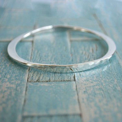 A silver bangle bracelet with a hammered texture and polished finish, shown on its side with a woven blue background. 