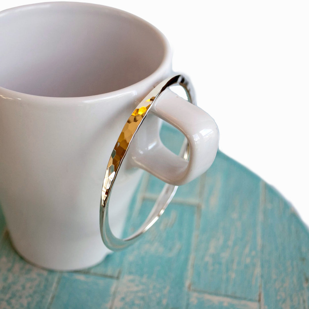 A silver bangle bracelet with a hammered texture and polished finish, displayed on the handle of a white mug.