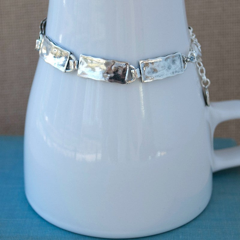 A sterling silver bracelet with links of slightly hammered texture is displayed around a white ceramic cup.