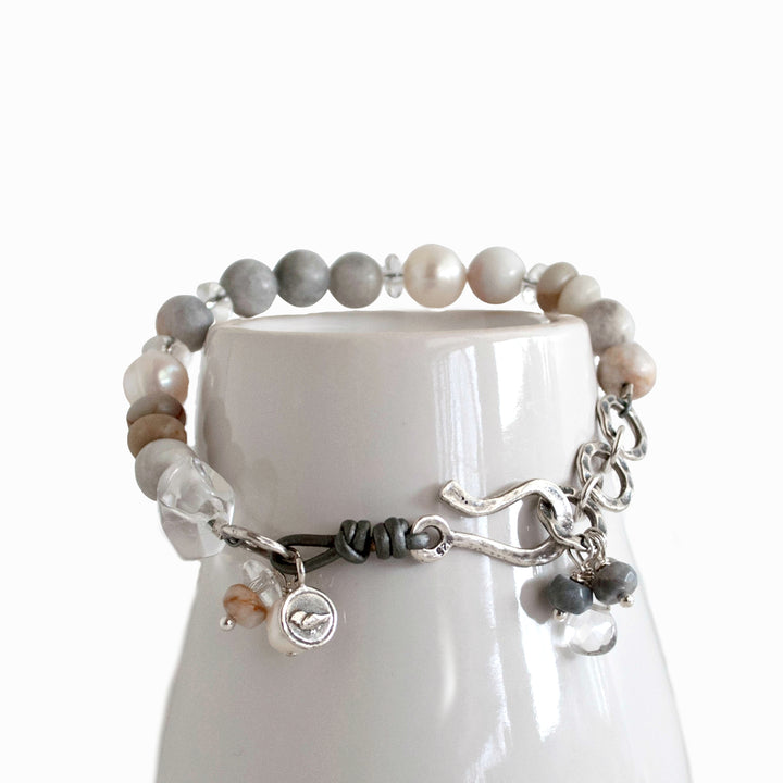 A bracelet with a mix of gray and white beads, pearls, and clear quartz, featuring a sterling silver clasp and leather detailing, is displayed on a white ceramic piece.
