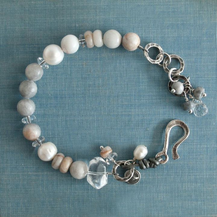 A bracelet with a mix of gray and white beads, pearls, and clear quartz, featuring a sterling silver clasp and leather detailing, is displayed on a blue linen background