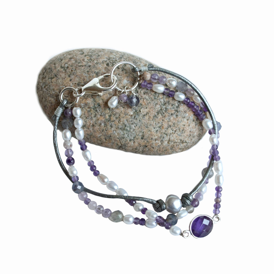 A mixed gemstone bracelet with tiers of freshwater pearls, amethyst, and iolite, featuring leather and a sterling lobster clasp, displayed on a rock.