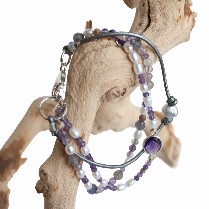 A mixed gemstone bracelet with tiers of freshwater pearls, amethyst, and iolite, featuring leather and a sterling lobster clasp, displayed on driftwood.