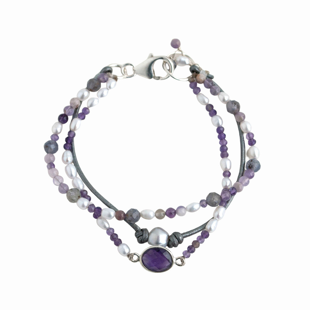 A mixed gemstone bracelet with tiers of freshwater pearls, amethyst, and iolite, featuring leather and a sterling lobster clasp, shown from above with a white background. 