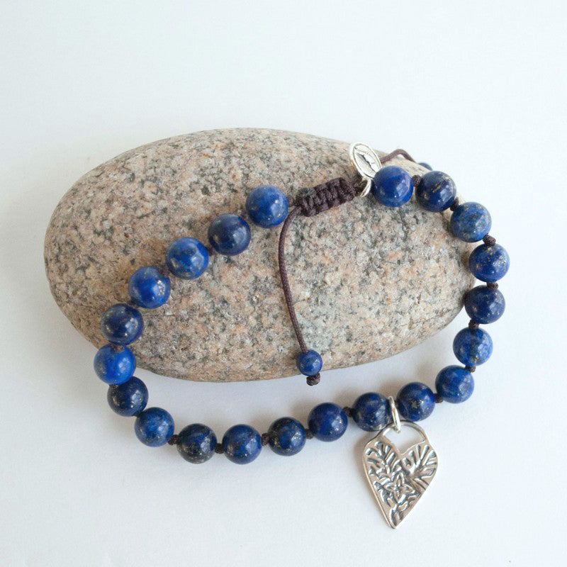 Bracelet consisting of smooth round 1/4" lapis beads on a brown adjustable silk cord. Features a sterling silver heart dangle. Shown on a rock with white background. 