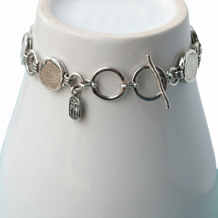 A sterling silver link bracelet with a textured finish and an adjustable toggle closure. Close up shown on white pottery.