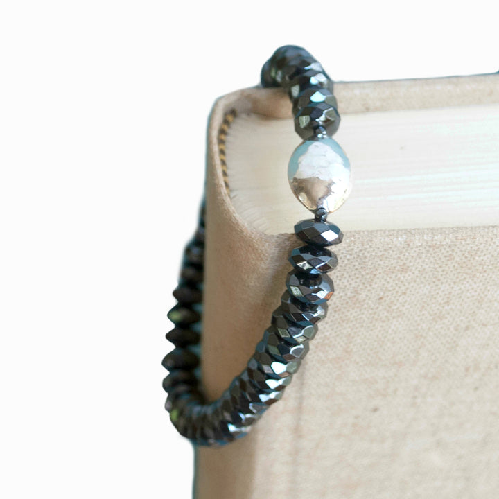 A stretch bracelet with faceted hematite beads and a hammered sterling silver centerpiece bead, displayed on a linen book.