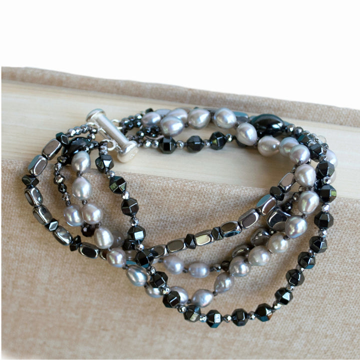 Multi-strand bracelet- crafted with grey freshwater pearl, a variety of hematite, and crystal. Finished witha  .925 sterling silver bar clasp. Available in sizes small, medium, and large. Shown on a linen book.