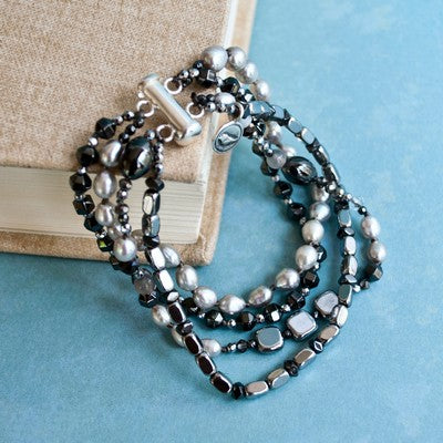 Multi-strand bracelet- crafted with grey freshwater pearl, a variety of hematite, and crystal. Finished with a .925 sterling silver bar clasp. Available in sizes small, medium, and large. Shown in an alternative view on a linen book. 