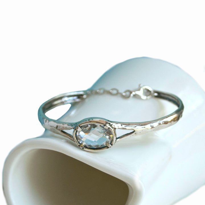 Silver Bracelet with a beautiful oval faceted clear CZ. Cuff-style bracelet with a safety chain. Artisan-crafted sterling silver bracelet shown on a white ceramic piece.