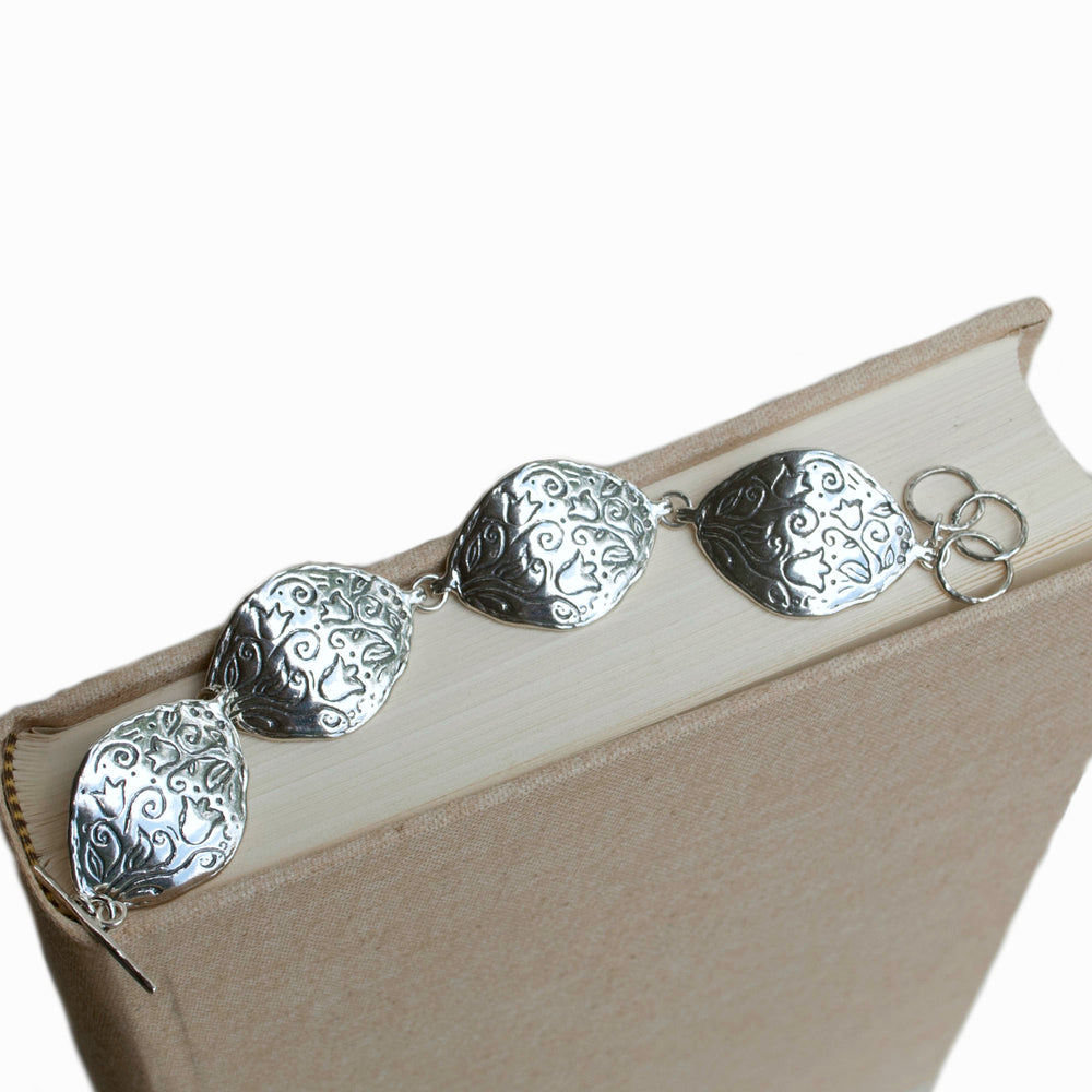 A sterling silver bracelet with a floral pattern, featuring four curved pieces and an adjustable toggle clasp. Shown on the spine of a linen book. 