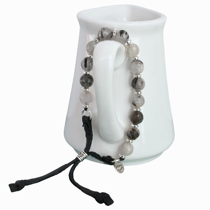 Stone Bracelet on genuine leather with a slide bead. Sterling silver accents and black rutilated quartz. Shown hanging on a small pitcher with a white background. 