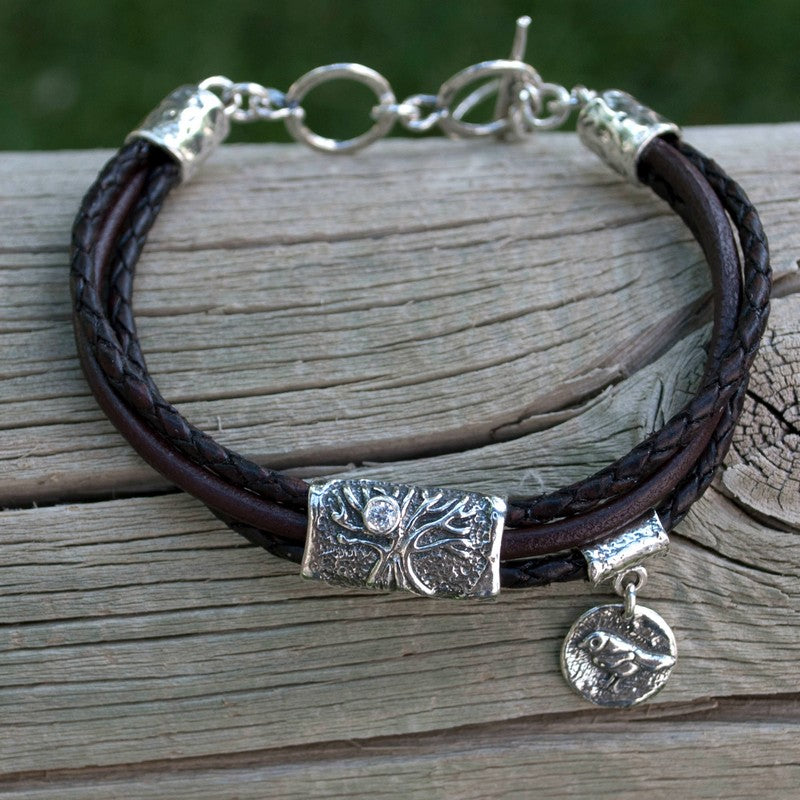A brown multi-strand leather bracelet with a silver Tree of Life charm and a bezel-set clear stone, displayed on a wood piece. 