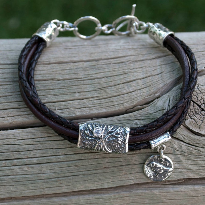 A brown multi-strand leather bracelet with a silver Tree of Life charm and a bezel-set clear stone, displayed on a wood piece. 