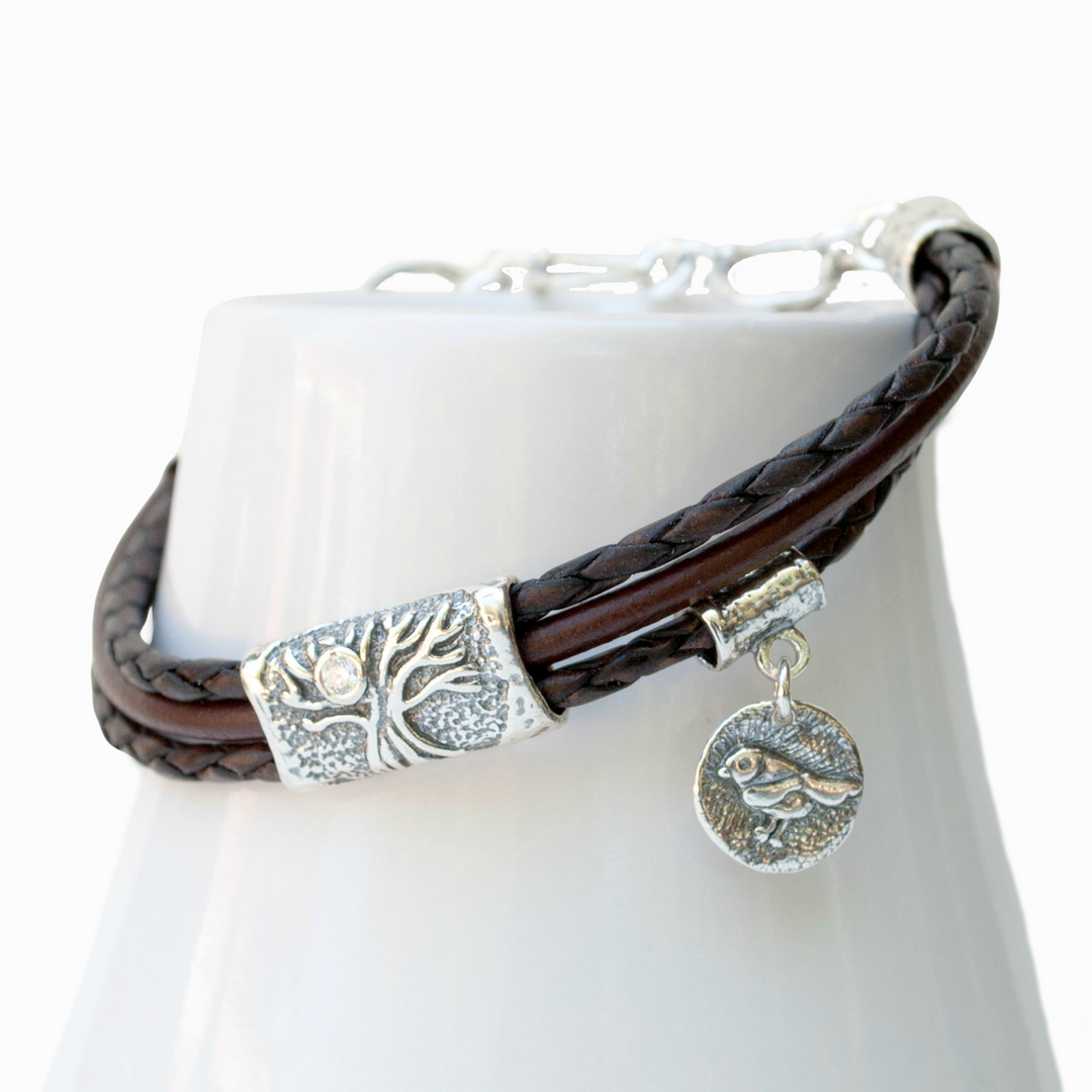 A brown multi-strand leather bracelet with a silver Tree of Life charm and a bezel-set clear stone, displayed on a white ceramic piece.