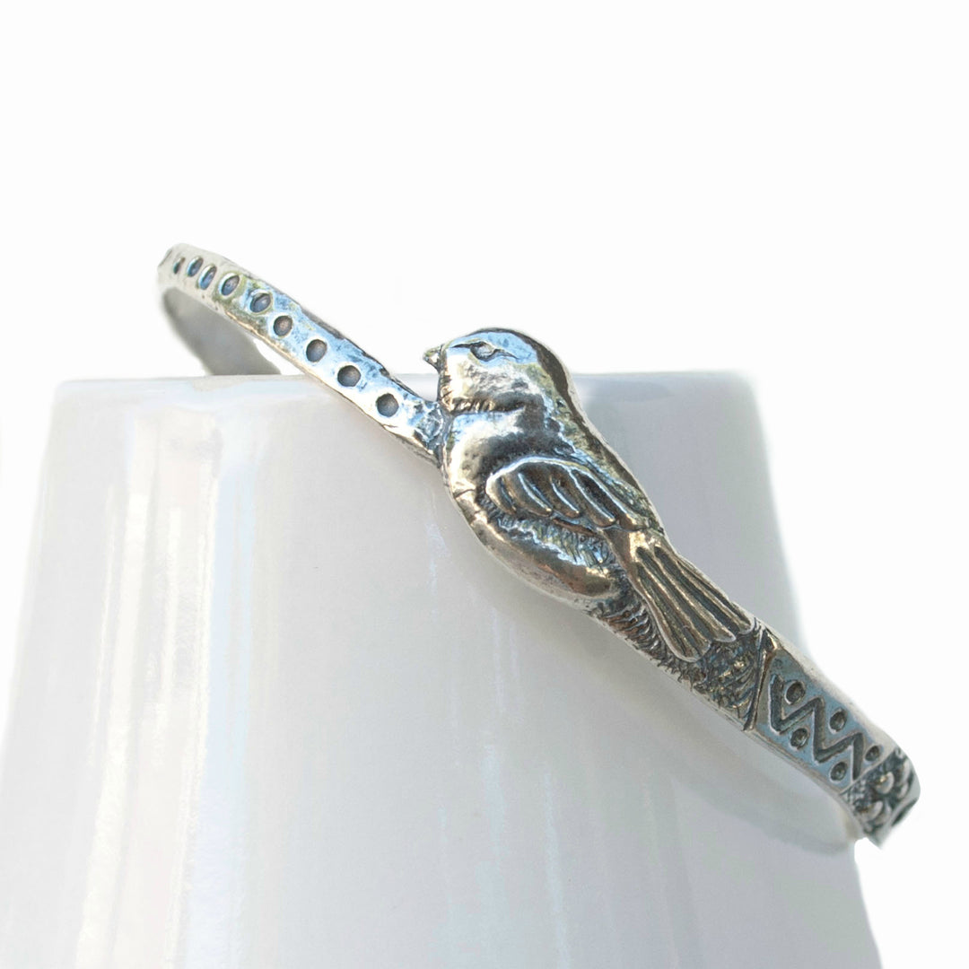 Silver Bangle Bracelet - .925 sterling silver crafted using the lost wax casting technique. Silver bangle bracelet featuring a textured pattern and a bird. Front of Bracelet shown on a white ceramic piece.
