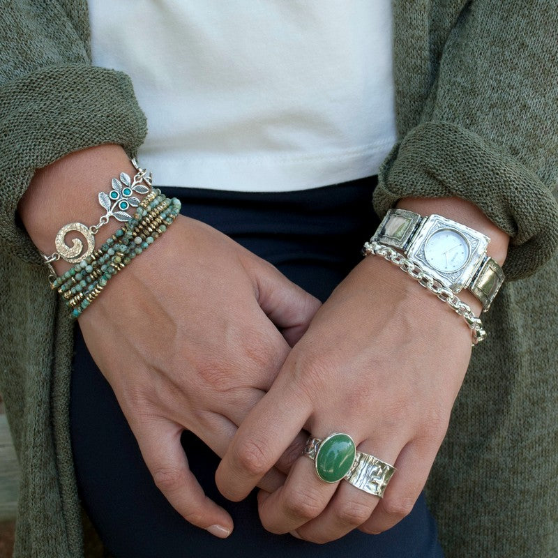A sterling silver charm bracelet with a variety of linked chains and charms, including a turquoise stone and a green adventurine, with an adjustable toggle closure. Shown on a model.