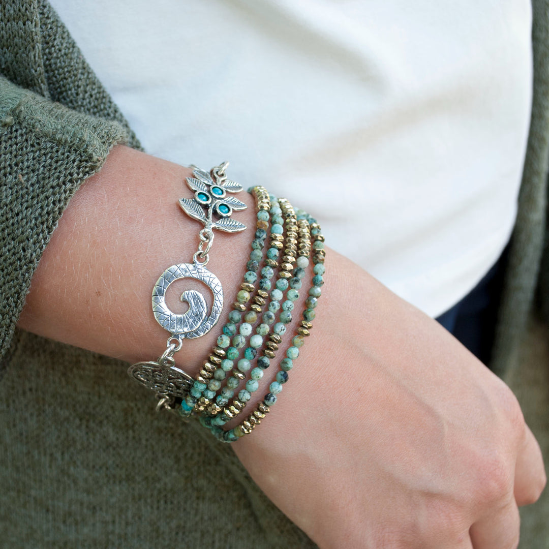 A sterling silver charm bracelet with a variety of linked chains and charms, including a turquoise stone and a green adventurine, with an adjustable toggle closure. Shown on a model&