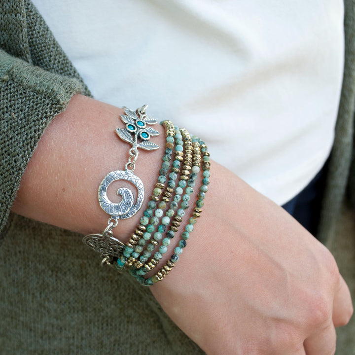 A sterling silver charm bracelet with a variety of linked chains and charms, including a turquoise stone and a green adventurine, with an adjustable toggle closure. Shown on a model&