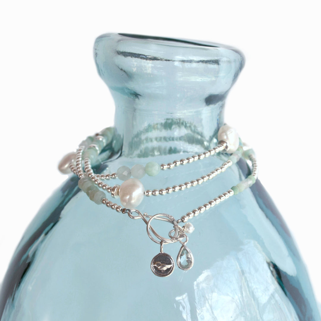 Convertible Necklace- wear as a wrap bracelet or necklace. Crafted of white freshwater pearls, aquamarine, amazonite and .925 sterling silver. Shown as a bracelet on an aqua blue vase.