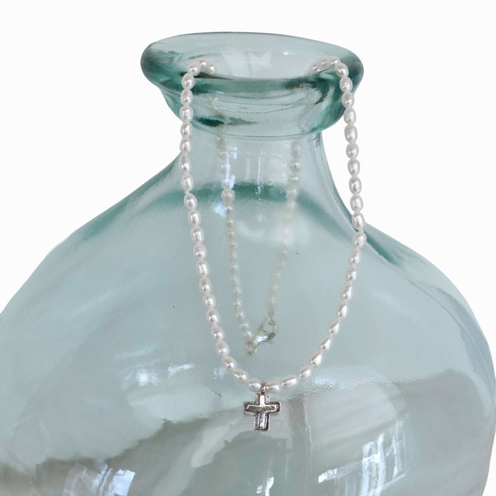 A necklace with a raised, textured sterling silver cross centered among luminous, freshwater seed pearls is shown on hanging on an aqua glass bottle. 