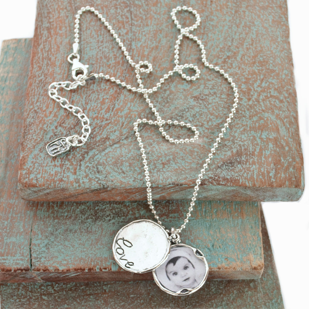 A sterling silver locket necklace with a bead chain and a locket etched with the word 'love', featuring a small photo peeking out from the front. Shown on wood blocks with a white background.