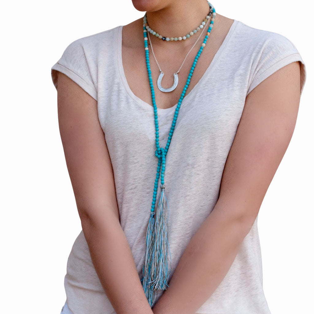 A model wearing a white sleeveless top and white pants, adorned with a long beaded necklace featuring matte amazonite and turquoise, along with matching earrings and bracelets. The necklace is shown doubled as a choker and then knotted. 