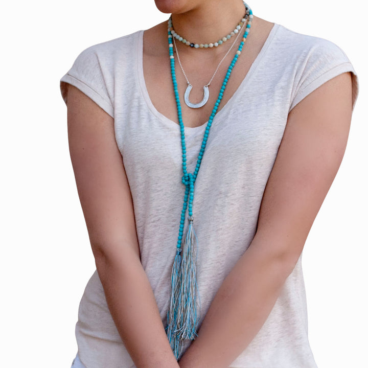 A model wearing a white sleeveless top and white pants, adorned with a long beaded necklace featuring matte amazonite and turquoise, along with matching earrings and bracelets. The necklace is shown doubled as a choker and then knotted. 