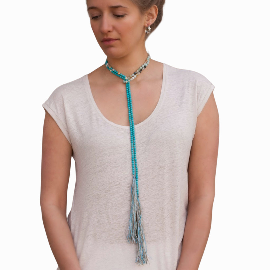 A model wearing a white sleeveless top and white pants, adorned with a long beaded necklace featuring matte amazonite and turquoise, along with matching earrings and bracelets. The necklace is shown worn as a lariat.