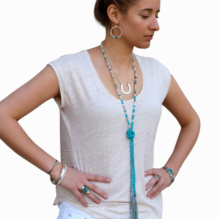 A model wearing a white sleeveless top and white pants, adorned with a long beaded necklace featuring matte amazonite and turquoise, along with matching earrings and bracelets. The necklace is shown long and knotted. 