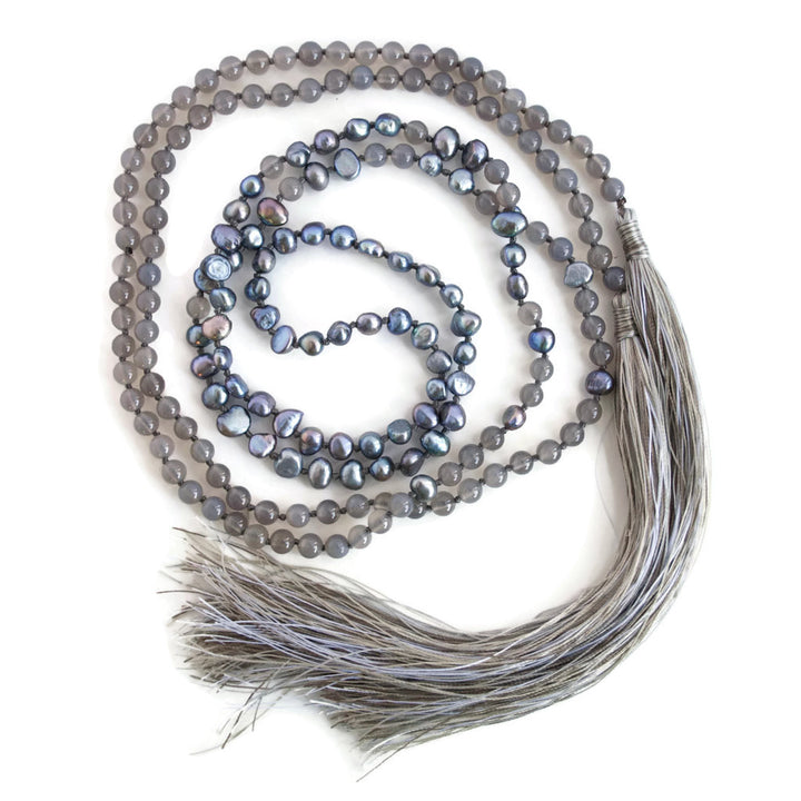 A lariat necklace with peacock freshwater pearls, grey agate, and grey cord, featuring long variegated tassels, is shown wrapped up in full on a white background. 