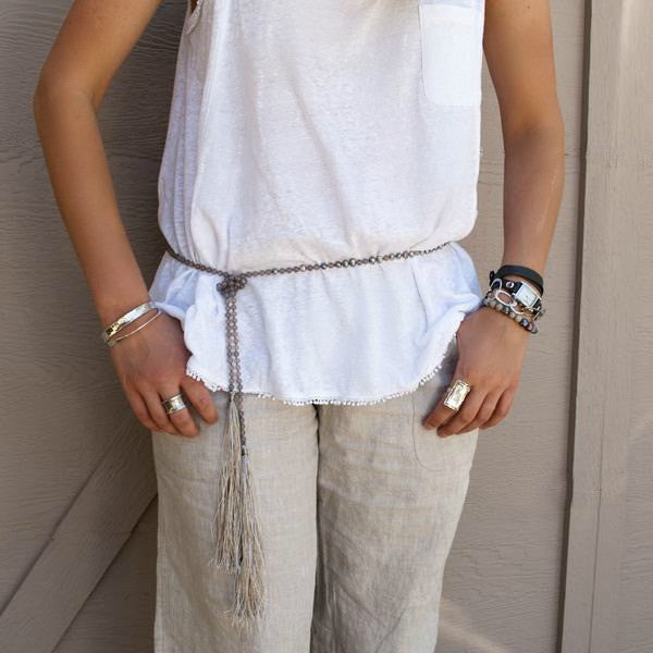 A lariat necklace with peacock freshwater pearls, grey agate, and grey cord, featuring long variegated tassels, is shown worn as a belt with a white shirt and tan linen pants. 