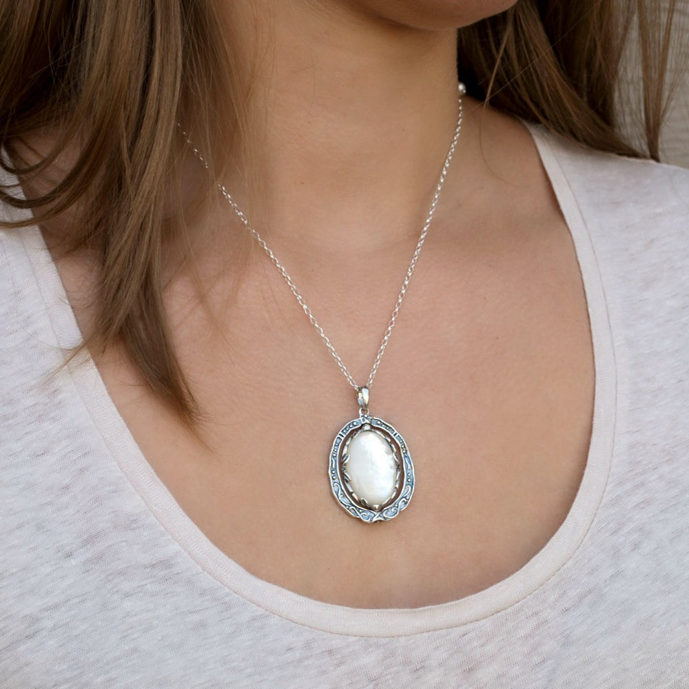 A sterling silver reversible necklace pendant with a filigree cross and mother-of-pearl on one side, and a plain mother-of-pearl on the reversible side, attached to a silver chain. Shown on a model with the mother-of-pearl side showing. 