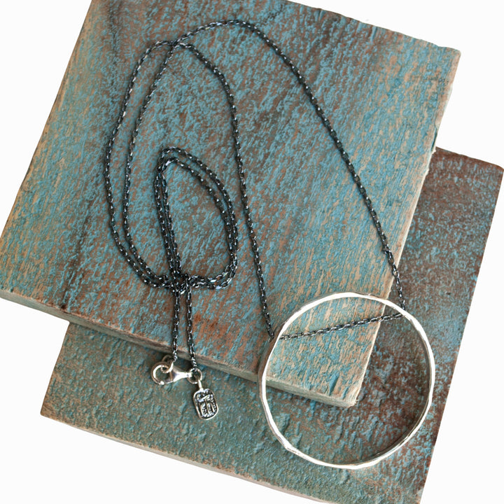 Long necklace with an oxidized sterling silver chain holds an organic circle of sterling silver. Shown on wooden tiles. 